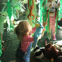 Mała dziewczynka bawi się porozwieszanymi zabawkami. Pomieszczenie przypomina dżunglę, w której wiszą liany i zwierzęta.
