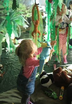 Mała dziewczynka bawi się porozwieszanymi zabawkami. Pomieszczenie przypomina dżunglę, w której wiszą liany i zwierzęta.