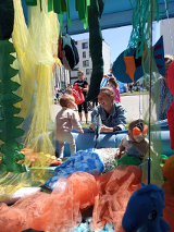 Małe dzieci siedzą w instalacji wielozmysłowej i bawią się zabawkami. Obok klęka mama jednego z dziecka i śmieje się do niego. Za nimi inne dzieci z rodzicami.