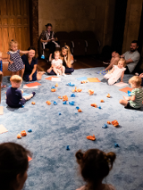 Zdjęcie sali, w której na podlodze pokrytej jasno niebieskim materiałem siedzą dzieci, przy nich rodzice. Na środku rozrzucone niebieskie i pomarańczowe zgniecione kawałki papieru.
