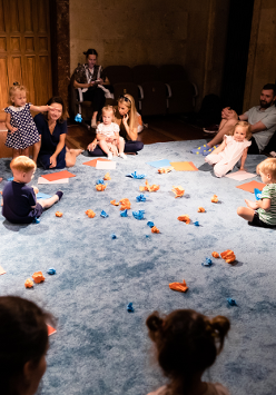 Zdjęcie sali, w której na podlodze pokrytej jasno niebieskim materiałem siedzą dzieci, przy nich rodzice. Na środku rozrzucone niebieskie i pomarańczowe zgniecione kawałki papieru.