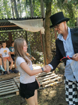 zdjęcie dziewczynki podającej rękę mężczyźnie we fraku i kapeluszu, w tle dzieci siedzące w altanie