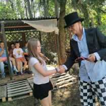 zdjęcie dziewczynki podającej rękę mężczyźnie we fraku i kapeluszu, w tle dzieci siedzące w altanie