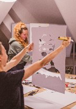 Trzy kobiety na warsztatach plastycznych. Jedna ogląda portret, dwie stoją przy stole z rozłożonymi materiałami plastycznymi (pędzle, kartki, farby).