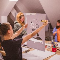 Trzy kobiety na warsztatach plastycznych. Jedna ogląda portret, dwie stoją przy stole z rozłożonymi materiałami plastycznymi (pędzle, kartki, farby).