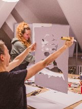 Trzy kobiety na warsztatach plastycznych. Jedna ogląda portret, dwie stoją przy stole z rozłożonymi materiałami plastycznymi (pędzle, kartki, farby).