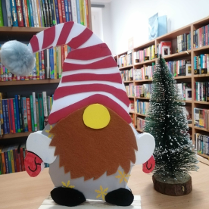 Na pierwszym planie świąteczny skrzat z brodą i czerwoną czapeczką. Obok ozdobna choinka. W tle regały z książkami.