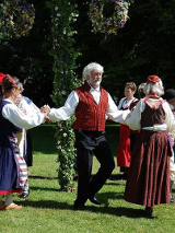 Na zdjęciu widać grupę ludzi, głównie kobiet, ubranych w tradycyjne skandynawskie stroje ludowe. Tańczą w kole wokół maja w parku. Kobiety noszą jaskrawo kolorowe suknie z misterną haftowaną ozdobą, a mężczyźni mają białe koszule z kamizelkami. Maj jest udekorowany kwiatami, wstążkami i zielenią. Scena wydaje się być uroczysta i radosna.