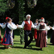 Na zdjęciu widać grupę ludzi, głównie kobiet, ubranych w tradycyjne skandynawskie stroje ludowe. Tańczą w kole wokół maja w parku. Kobiety noszą jaskrawo kolorowe suknie z misterną haftowaną ozdobą, a mężczyźni mają białe koszule z kamizelkami. Maj jest udekorowany kwiatami, wstążkami i zielenią. Scena wydaje się być uroczysta i radosna.