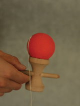 Zdjęcie, ręka trzyma drewniane urządzenie, do którego przymocowano za pomocą sznurka czerwoną piłkę.
