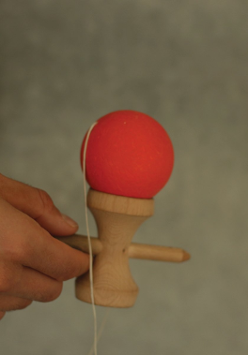 Zdjęcie, ręka trzyma drewniane urządzenie, do którego przymocowano za pomocą sznurka czerwoną piłkę.