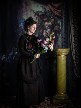 Zdjęcie, na ciemnym tle stoi kobieta w długiej sukni, trzymająca bujkiet kwiatów.