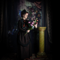 Zdjęcie, na ciemnym tle stoi kobieta w długiej sukni, trzymająca bujkiet kwiatów.