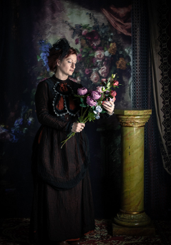 Zdjęcie, na ciemnym tle stoi kobieta w długiej sukni, trzymająca bujkiet kwiatów.