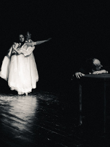 Na scenie widoczny mężczyzna opierający głowę i ręce o stolik, w tle stoją dwie kobiety w długich białych sukniach, unoszą ręce do góry