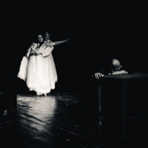 Na scenie widoczny mężczyzna opierający głowę i ręce o stolik, w tle stoją dwie kobiety w długich białych sukniach, unoszą ręce do góry