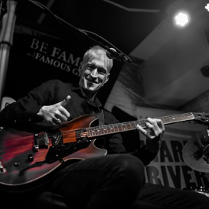 Mężczyzna na scenie pokazuje gest kciuka w górę grając na gitarze.