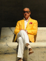 Starszy mężczyzna w białych spodniach, koszuli i zółtek marynarce. W kieszeni ma czerwoną chusteczkę. Na nosie czarne okulary. Siedzi na marmurowej ławce.