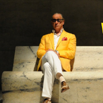 Starszy mężczyzna w białych spodniach, koszuli i zółtek marynarce. W kieszeni ma czerwoną chusteczkę. Na nosie czarne okulary. Siedzi na marmurowej ławce.