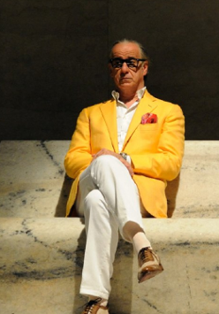 Starszy mężczyzna w białych spodniach, koszuli i zółtek marynarce. W kieszeni ma czerwoną chusteczkę. Na nosie czarne okulary. Siedzi na marmurowej ławce.