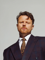 Artysta w garniturze i krawacie na tle szarej ściany.