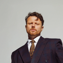 Artysta w garniturze i krawacie na tle szarej ściany.