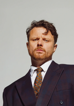 Artysta w garniturze i krawacie na tle szarej ściany.