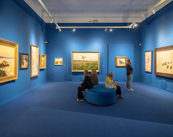 Wnętrze muzeum, niebieskie ściany, na nich obrazy, publiczność ogląda stojąc i siedząc