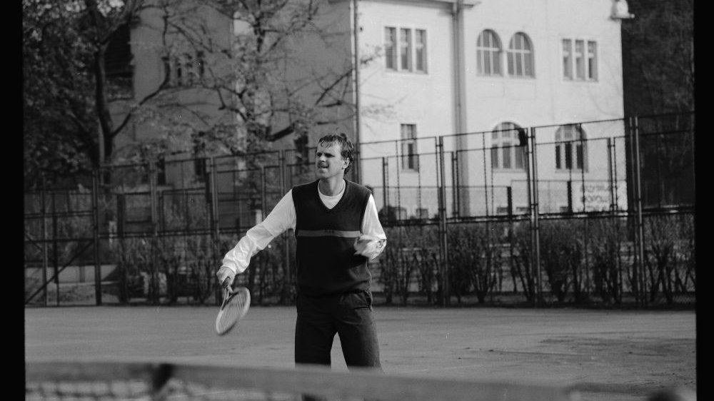 Czarno-biała fotografia mężczyzny, który gra w tenisa. Za nim duży biały budynek.