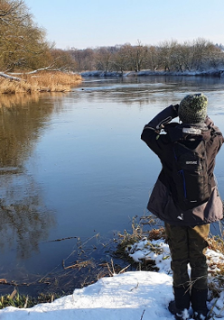 Osoba stoi nad brzegiem rzeki w zimowej aurze, nad jej brzegami nachylają się drzewa.