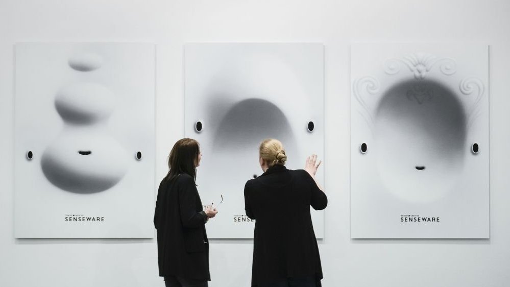 Dwie kobiety w czarnych strojach stoją tyłem. Patrzą na trzy duże obrazy zawieszone na ścianie. Jedna z nich trzyma okulary, druga gestykuluje.