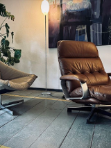 Zdjęcie przedstawia przytulny kącik wewnętrzny, który można znaleźć w domu, biurze lub salonie. Znajdują się w nim dwa różne rodzaje krzeseł ustawionych naprzeciwko siebie, tworząc przestrzeń do rozmów. Po lewej stronie znajduje się krzesło w nowoczesnym stylu z jasnoszarą tapicerką z tkaniny i minimalistyczną metalową ramą. Po prawej stronie znajduje się pluszowa skórzana leżanka w bogatym brązowym kolorze z drewnianymi akcentami na podłokietnikach, co sugeruje wygodę i relaks.