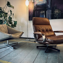 Zdjęcie przedstawia przytulny kącik wewnętrzny, który można znaleźć w domu, biurze lub salonie. Znajdują się w nim dwa różne rodzaje krzeseł ustawionych naprzeciwko siebie, tworząc przestrzeń do rozmów. Po lewej stronie znajduje się krzesło w nowoczesnym stylu z jasnoszarą tapicerką z tkaniny i minimalistyczną metalową ramą. Po prawej stronie znajduje się pluszowa skórzana leżanka w bogatym brązowym kolorze z drewnianymi akcentami na podłokietnikach, co sugeruje wygodę i relaks.