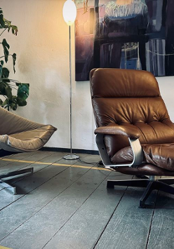 Zdjęcie przedstawia przytulny kącik wewnętrzny, który można znaleźć w domu, biurze lub salonie. Znajdują się w nim dwa różne rodzaje krzeseł ustawionych naprzeciwko siebie, tworząc przestrzeń do rozmów. Po lewej stronie znajduje się krzesło w nowoczesnym stylu z jasnoszarą tapicerką z tkaniny i minimalistyczną metalową ramą. Po prawej stronie znajduje się pluszowa skórzana leżanka w bogatym brązowym kolorze z drewnianymi akcentami na podłokietnikach, co sugeruje wygodę i relaks.