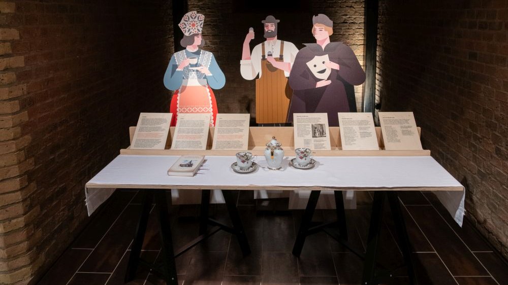 Papierowe postaci bamberki, rzemieślnika i aktora stoją za stołem z filiżankami i dokumentami.