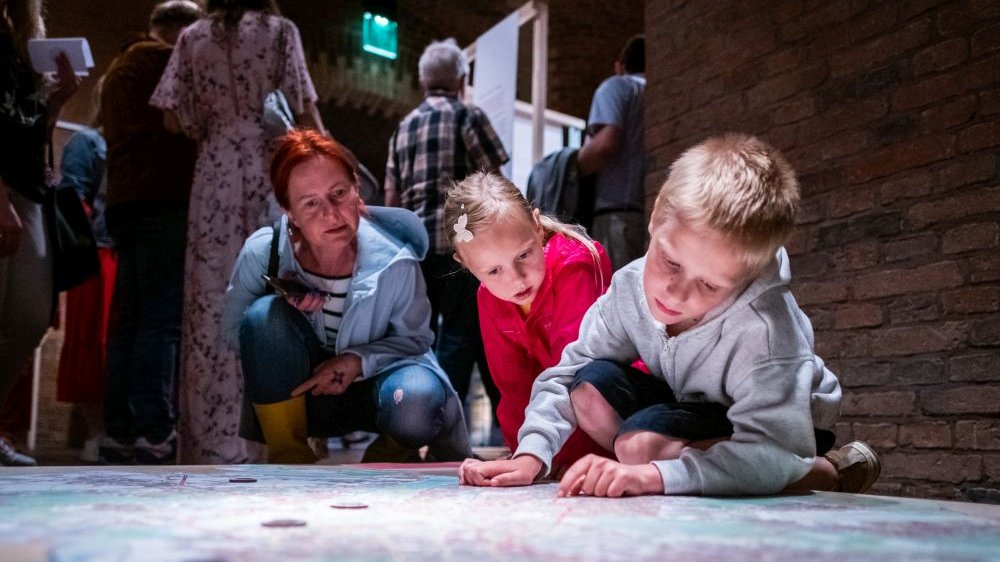 Zwiedzający wystawę w Galerii Śluza. Z przodu dwóje dzieci i kobieta czytając coś co jest napisane na podłodze.