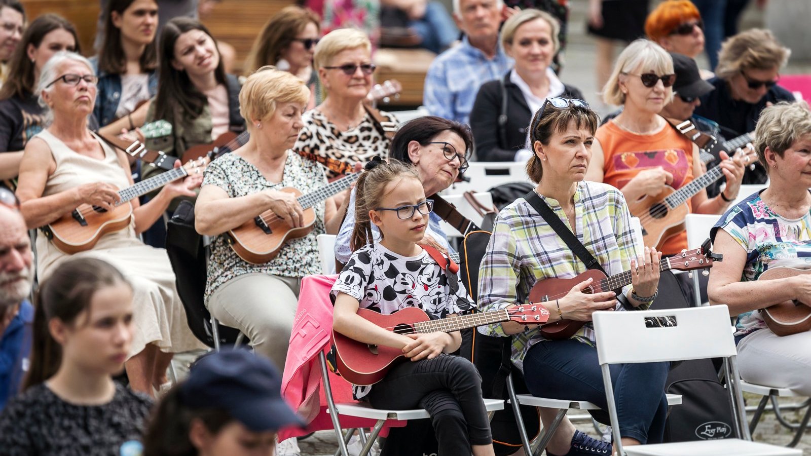 Zasłuchani i skupieni ludzie w różnym wieku i różnej płci siedzą na krzesłach na dziedzińcu Urzędu Miasta Poznania i grają na ukulele, instrumentach strunowych podobnych do małej gitary