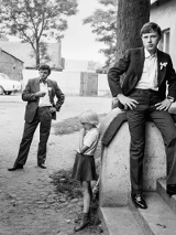 Czarnobiałe zdjęcie grupy młodych mężczyzn i dzieci. Mężczyźni ubrani są w retrro garnitury. Dzieci ubrane są w ciemne spodnie i jasne koszulki. Pozują na schodach pod budynkiem.