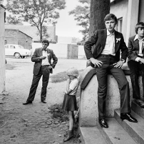 Czarnobiałe zdjęcie grupy młodych mężczyzn i dzieci. Mężczyźni ubrani są w retrro garnitury. Dzieci ubrane są w ciemne spodnie i jasne koszulki. Pozują na schodach pod budynkiem.