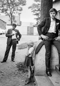 Czarnobiałe zdjęcie grupy młodych mężczyzn i dzieci. Mężczyźni ubrani są w retrro garnitury. Dzieci ubrane są w ciemne spodnie i jasne koszulki. Pozują na schodach pod budynkiem.