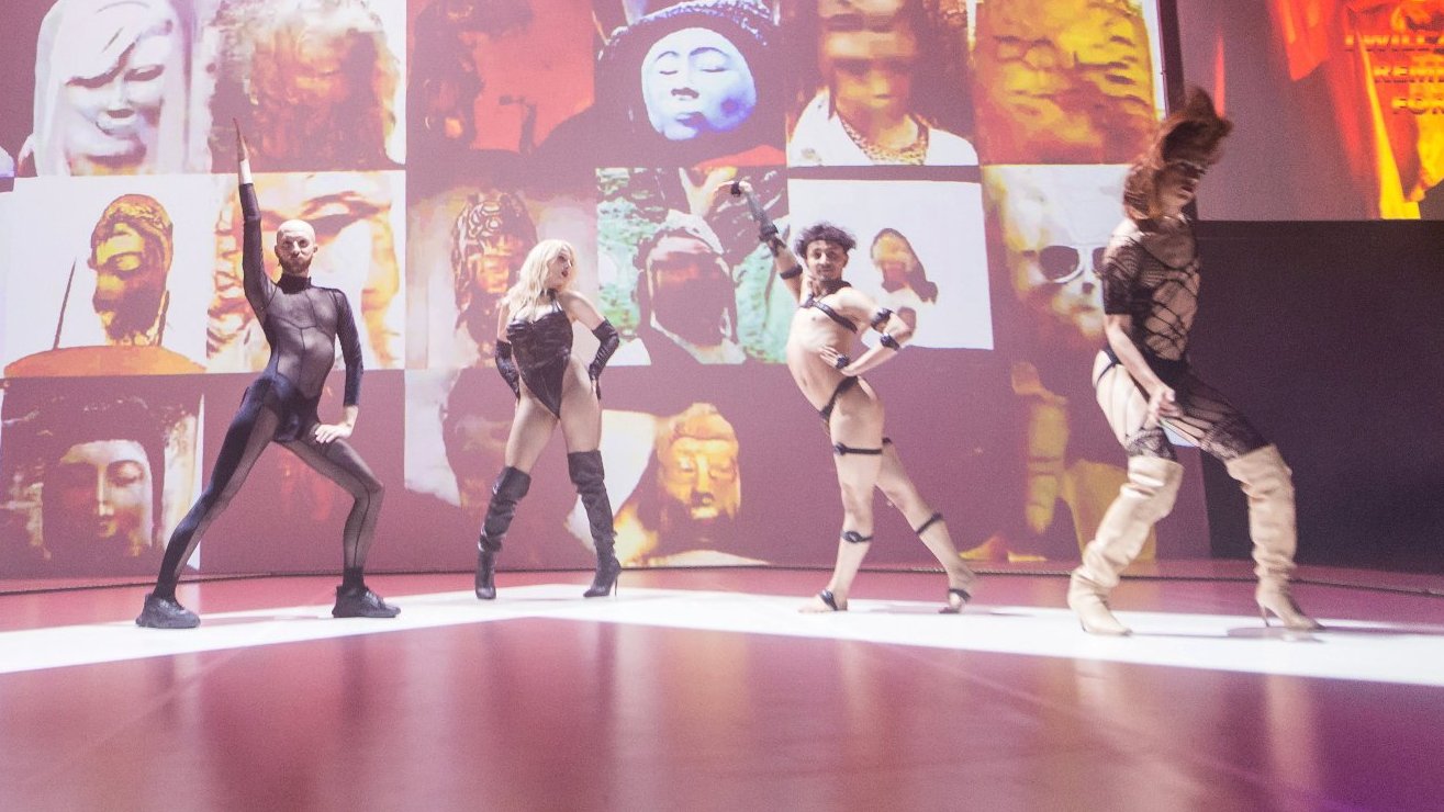 Pięć osób ze sceny voguingowej występuje na scenie. W tle kolorowa grafika z twarzami bóstw.