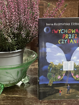 Zdjęcie, obok doniczki z wrzosami stoi książka, pt."Wychowanie przez czytanie", Irena Koźmińska, Elżbieta Olszewska. Na okładce znajduje się otwarta książka, z której wyrastają ogromne skrzydła motyla. Do ksiązki prowadzi drabina, przed którą stoi dziecko i dorosły mężczyzna..