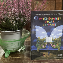 Zdjęcie, obok doniczki z wrzosami stoi książka, pt."Wychowanie przez czytanie", Irena Koźmińska, Elżbieta Olszewska. Na okładce znajduje się otwarta książka, z której wyrastają ogromne skrzydła motyla. Do ksiązki prowadzi drabina, przed którą stoi dziecko i dorosły mężczyzna..