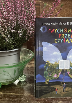 Zdjęcie, obok doniczki z wrzosami stoi książka, pt."Wychowanie przez czytanie", Irena Koźmińska, Elżbieta Olszewska. Na okładce znajduje się otwarta książka, z której wyrastają ogromne skrzydła motyla. Do ksiązki prowadzi drabina, przed którą stoi dziecko i dorosły mężczyzna..