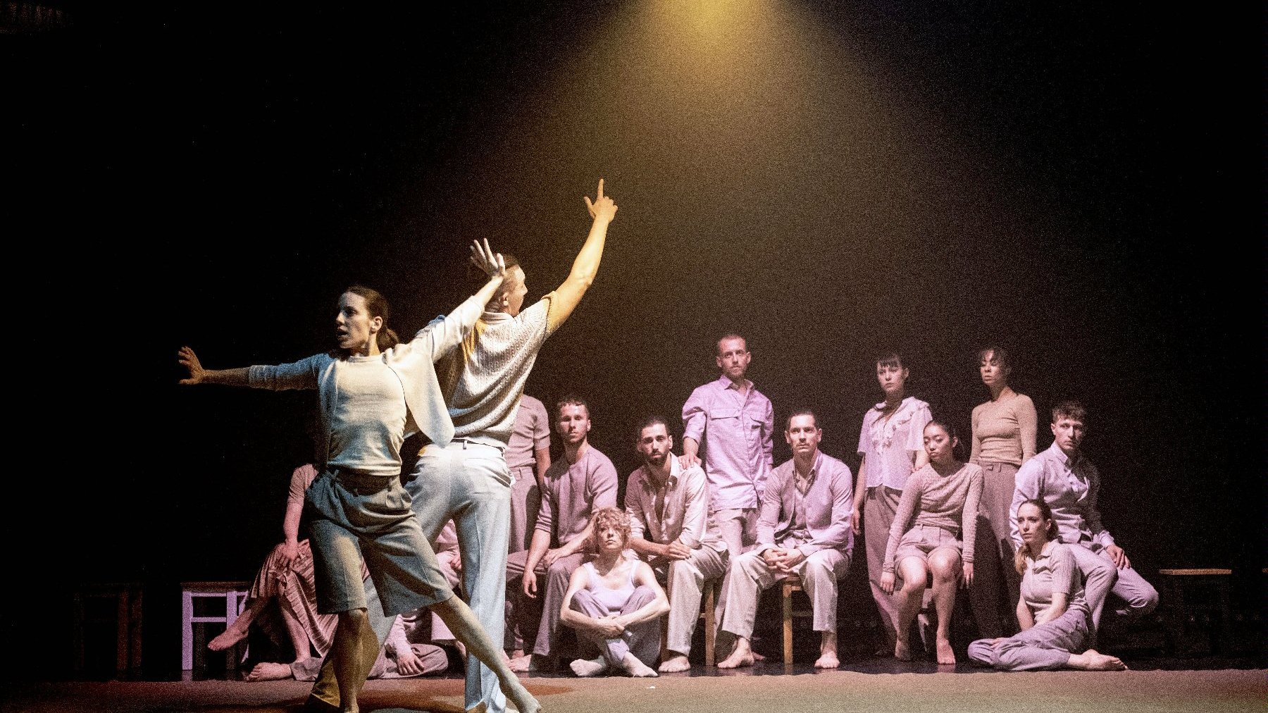 Mężczyzna i kobieta tańczą na środku sceny, inni tancerze siedzą przyglądając się im. Z góry oświetla ich lampa.