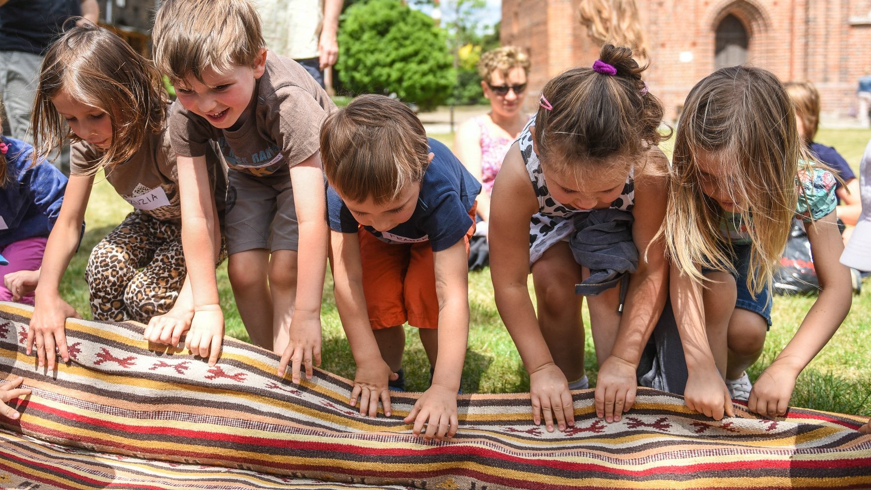 Dzieci bawią się na trawie, rozwijają dywan, w tle gotycki kościół