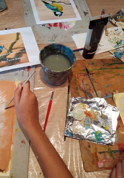 Farby, pędzle rozrzucone na stole. Miedzy nimi malunki kolorowych ptaktów.