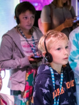 Grupa dzieci stoi, słuchając czegoś przez słuchawki.