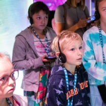 Grupa dzieci stoi, słuchając czegoś przez słuchawki.