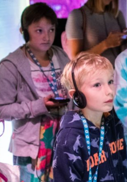 Grupa dzieci stoi, słuchając czegoś przez słuchawki.